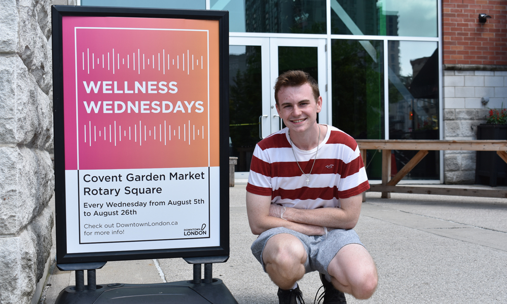 A photo of the website owner beside a white and salmon design at an event reading Wellness Wednesdays. Covent Garden Market Rotary Square. Every Wednesday from August 5th to August 26th. Check out DowntownLondon.ca for more info! This photo is meant to showcase my graphic design abilities.